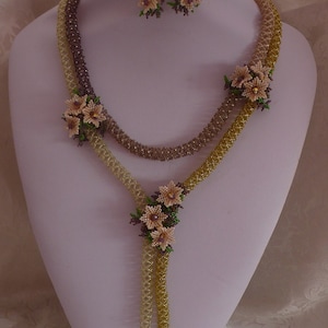 Può includere: Una delicata collana di perline dorate e marroni con un design floreale. La collana presenta un lungo ciondolo con un gruppo di piccoli fiori. Sono inclusi orecchini abbinati con un design di un singolo fiore.