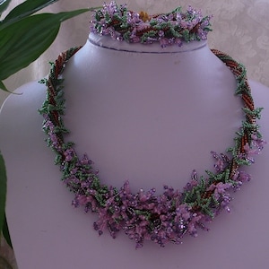 Può includere: Un delicato set di collana e braccialetto con perline, con un design floreale in perline viola, verdi e marroni. I gioielli sono esposti su un manichino bianco.