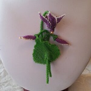 Può includere: Un fiore di perline viola con una foglia e uno stelo di perline verdi. Il fiore è fatto con perline di semi e ha una perla dorata al centro.