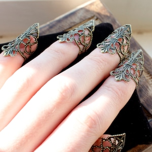 May include: Five gold-toned finger rings with a delicate, intricate design. The rings are worn on the fingers of a hand, with the tips of the rings pointing upwards.