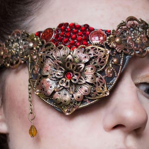 May include: A gold and red eye patch with a floral design and a dangling amber bead. The eye patch is worn by a person with brown hair and makeup.