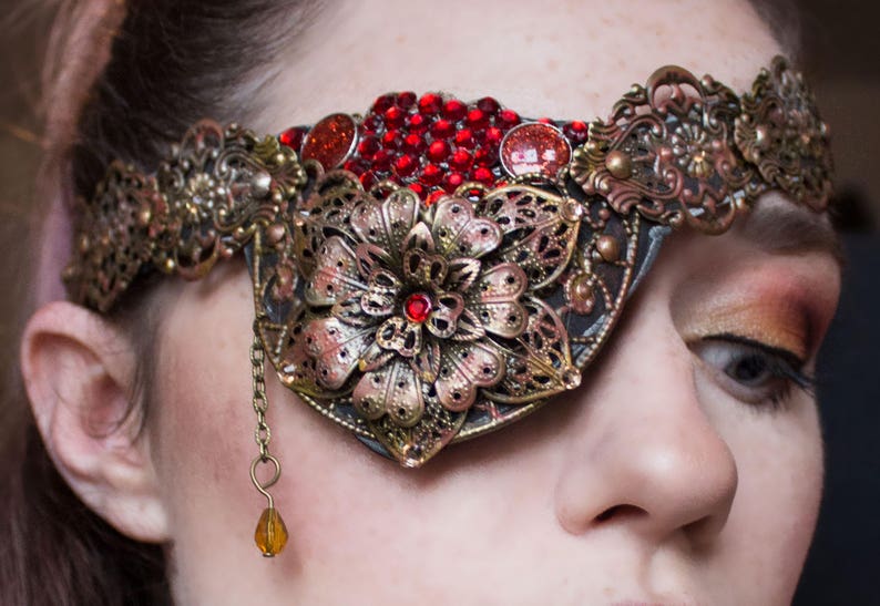 May include: A gold and red eye patch with a floral design and a dangling amber bead. The eye patch is worn by a person with brown hair and makeup.