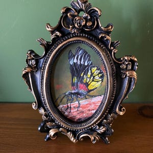 May include: A framed painting of a black, yellow, and red butterfly with white markings. The butterfly is perched on a brown rock. The frame is black with gold accents and an oval shape.