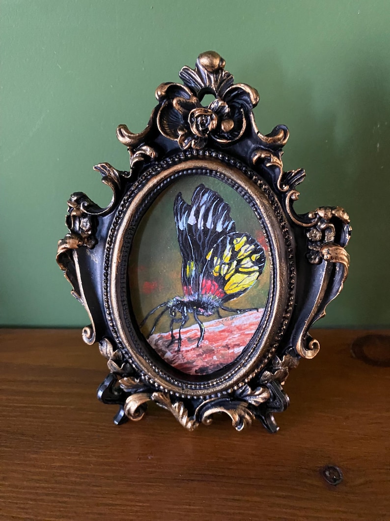 May include: A framed painting of a black, yellow, and red butterfly with white markings. The butterfly is perched on a brown rock. The frame is black with gold accents and an oval shape.