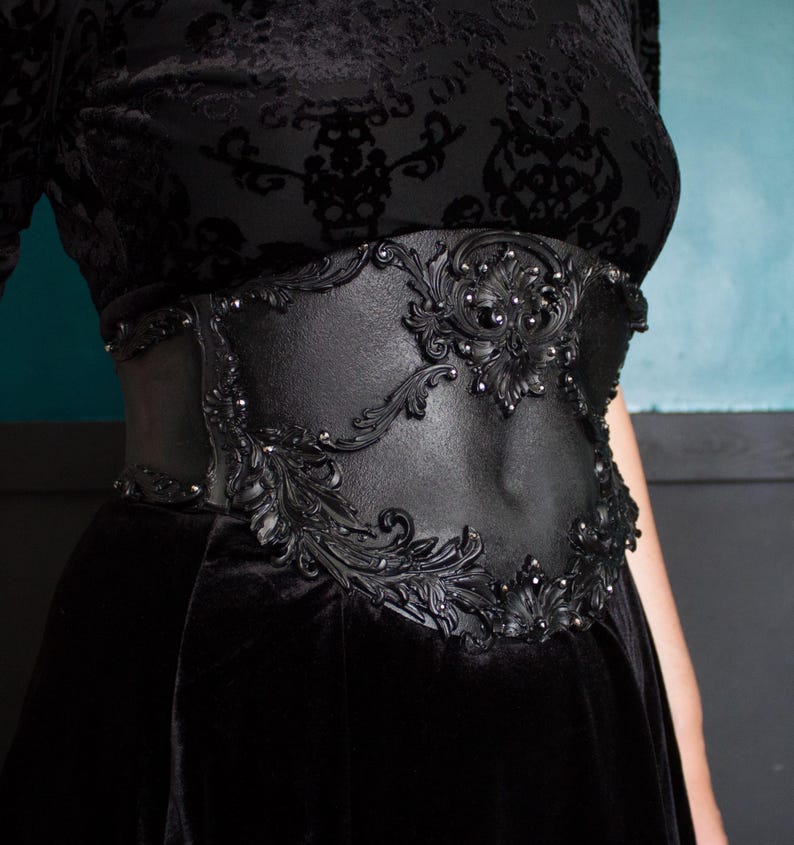 May include: Black corset belt with ornate, raised floral and scrollwork detailing. The belt features a central design with small, silver-colored embellishments. The belt is worn over a black velvet garment, creating a gothic or Victorian aesthetic.