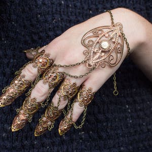 Puede incluir: Un conjunto de anillos de dedos adornados en tonos dorados con dise&ntilde;os intrincados y un solo strass transparente. Los anillos est&aacute;n conectados por cadenas y se llevan en varios dedos.