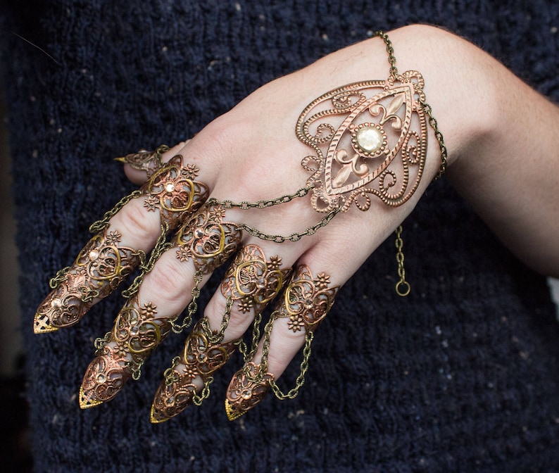 Puede incluir: Un conjunto de anillos de dedos adornados en tonos dorados con dise&ntilde;os intrincados y un solo strass transparente. Los anillos est&aacute;n conectados por cadenas y se llevan en varios dedos.