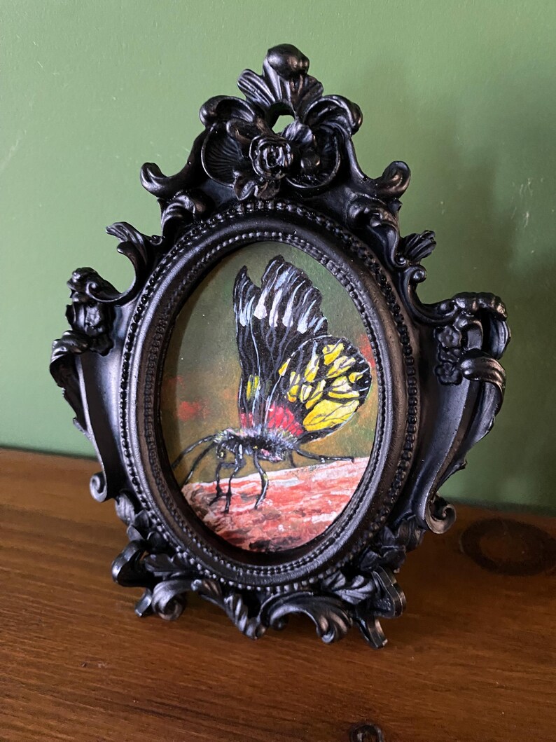 May include: A black and yellow butterfly with red accents is painted on a canvas in an oval-shaped frame. The frame is black and ornate with floral details.