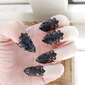 May include: Five black metal fingertip rings with a lacy design and blue accents. The rings are worn on a hand.