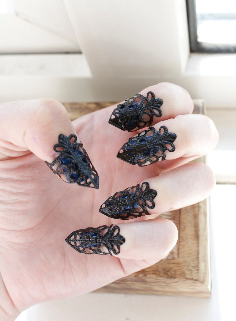 May include: Five black metal fingertip rings with a lacy design and blue accents. The rings are worn on a hand.