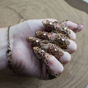 Puede incluir: Un conjunto de cinco anillos de dedo ornamentados y conectados, con un dise&ntilde;o de filigrana. Los anillos est&aacute;n hechos de lat&oacute;n y tienen un acabado de cobre. Cada anillo presenta una punta puntiaguda y est&aacute; adornado con diamantes de imitaci&oacute;n transparentes.