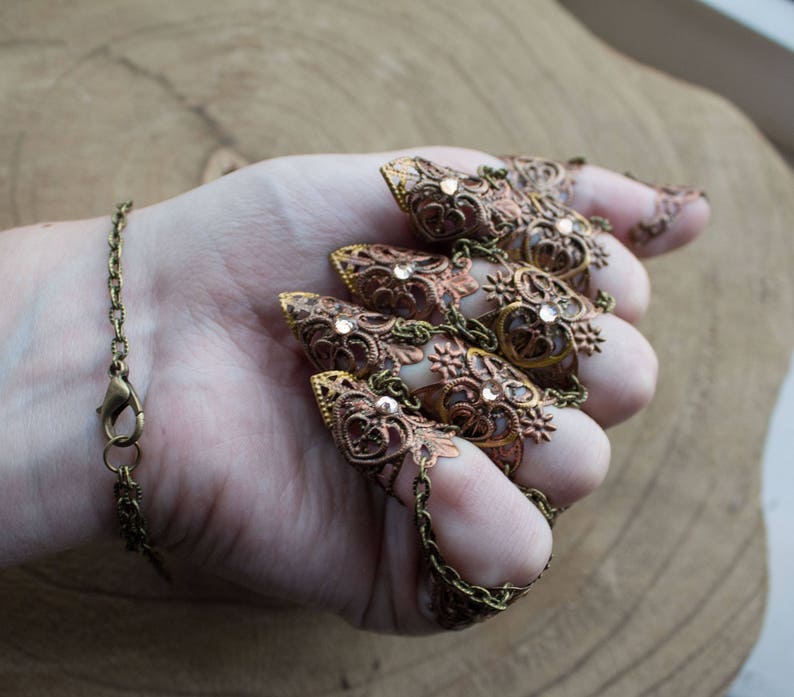 Puede incluir: Un conjunto de cinco anillos de dedo ornamentados y conectados, con un dise&ntilde;o de filigrana. Los anillos est&aacute;n hechos de lat&oacute;n y tienen un acabado de cobre. Cada anillo presenta una punta puntiaguda y est&aacute; adornado con diamantes de imitaci&oacute;n transparentes.