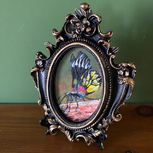 May include: A framed painting of a yellow and black butterfly with red accents, perched on a rock. The frame is black with gold accents and has an oval shape.