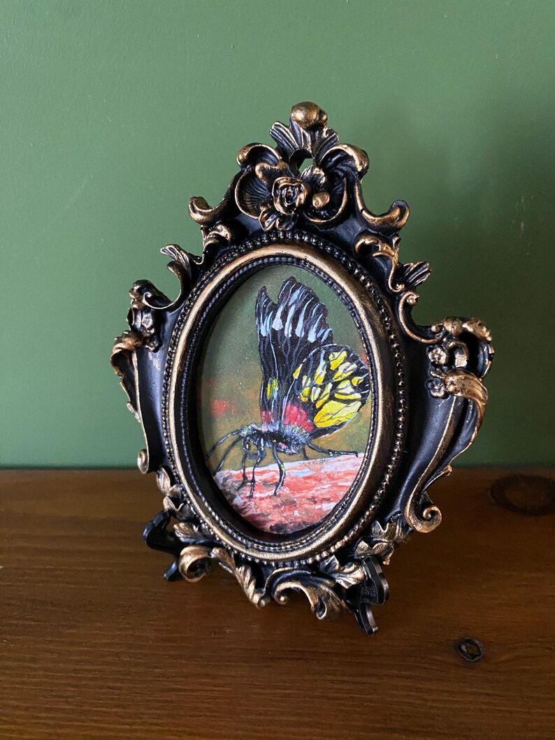 May include: A framed painting of a yellow and black butterfly with red accents, perched on a rock. The frame is black with gold accents and has an oval shape.