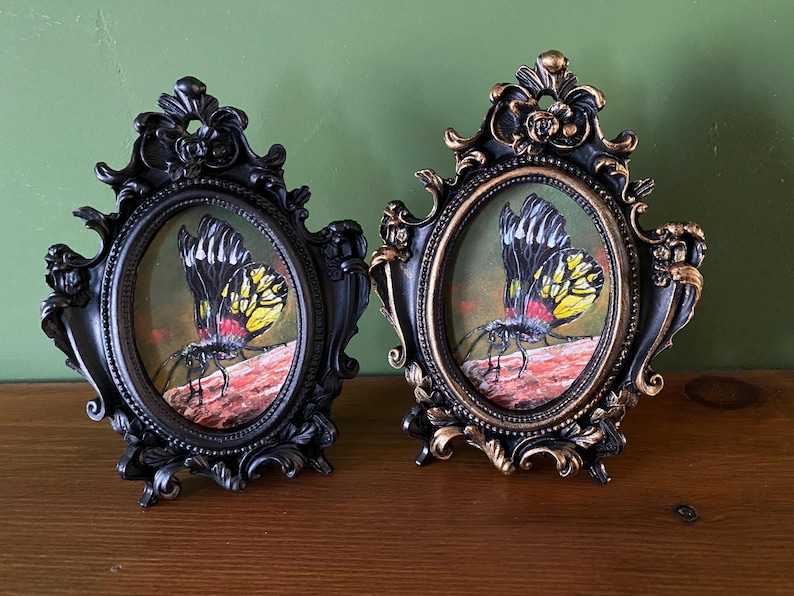 May include: Two ornate oval frames with black and gold finishes, each containing a painting of a yellow and black butterfly with red accents on a brown background.