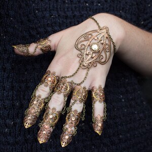Puede incluir: Un conjunto de anillos de dedo de color dorado y cobre con intrincados dise&ntilde;os florales. Los anillos est&aacute;n conectados por cadenas y presentan un gran colgante con un centro de perla.