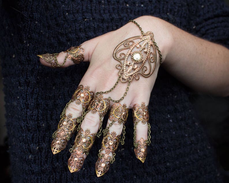 Puede incluir: Un conjunto de anillos de dedo de color dorado y cobre con intrincados dise&ntilde;os florales. Los anillos est&aacute;n conectados por cadenas y presentan un gran colgante con un centro de perla.