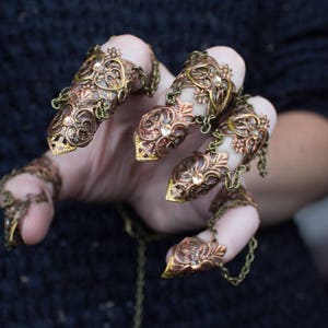 Puede incluir: Un conjunto de cinco anillos de dedo de oro y cobre con intrincados dise&ntilde;os florales. Los anillos est&aacute;n conectados por cadenas y se llevan en los dedos de una mano.