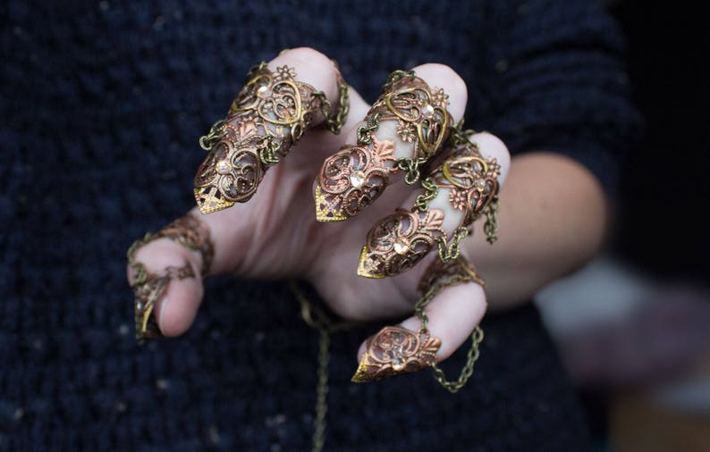 Puede incluir: Un conjunto de cinco anillos de dedo de oro y cobre con intrincados dise&ntilde;os florales. Los anillos est&aacute;n conectados por cadenas y se llevan en los dedos de una mano.