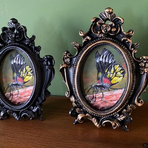 May include: Two ornate black and gold oval frames with a black background and gold trim. Each frame contains a painting of a yellow and black butterfly with red accents on a red and brown background.