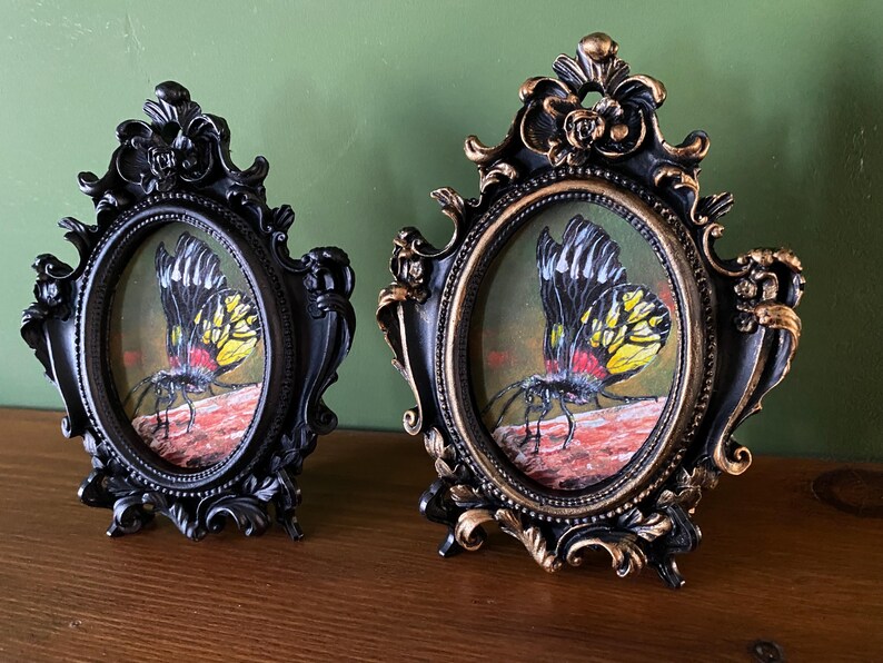 May include: Two ornate black and gold oval frames with a black background and gold trim. Each frame contains a painting of a yellow and black butterfly with red accents on a red and brown background.