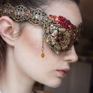 May include: A gold and red eye patch with a floral design and rhinestones. The eye patch is attached to a chain with a dangling amber bead.