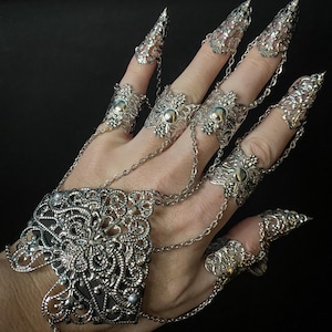 Könnte beinhalten: Ein silbernes Filigranarmband und Fingerringe mit krallenartigen Spitzen. Das Armband ist quadratisch und mit Ketten an die Ringe verbunden.