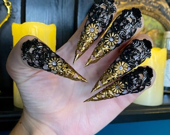 Puede incluir: Cinco anillos de uñas en forma de garra negros y dorados con intrincados diseños florales. Los anillos se llevan en una mano, con las puntas de las garras apuntando hacia afuera.