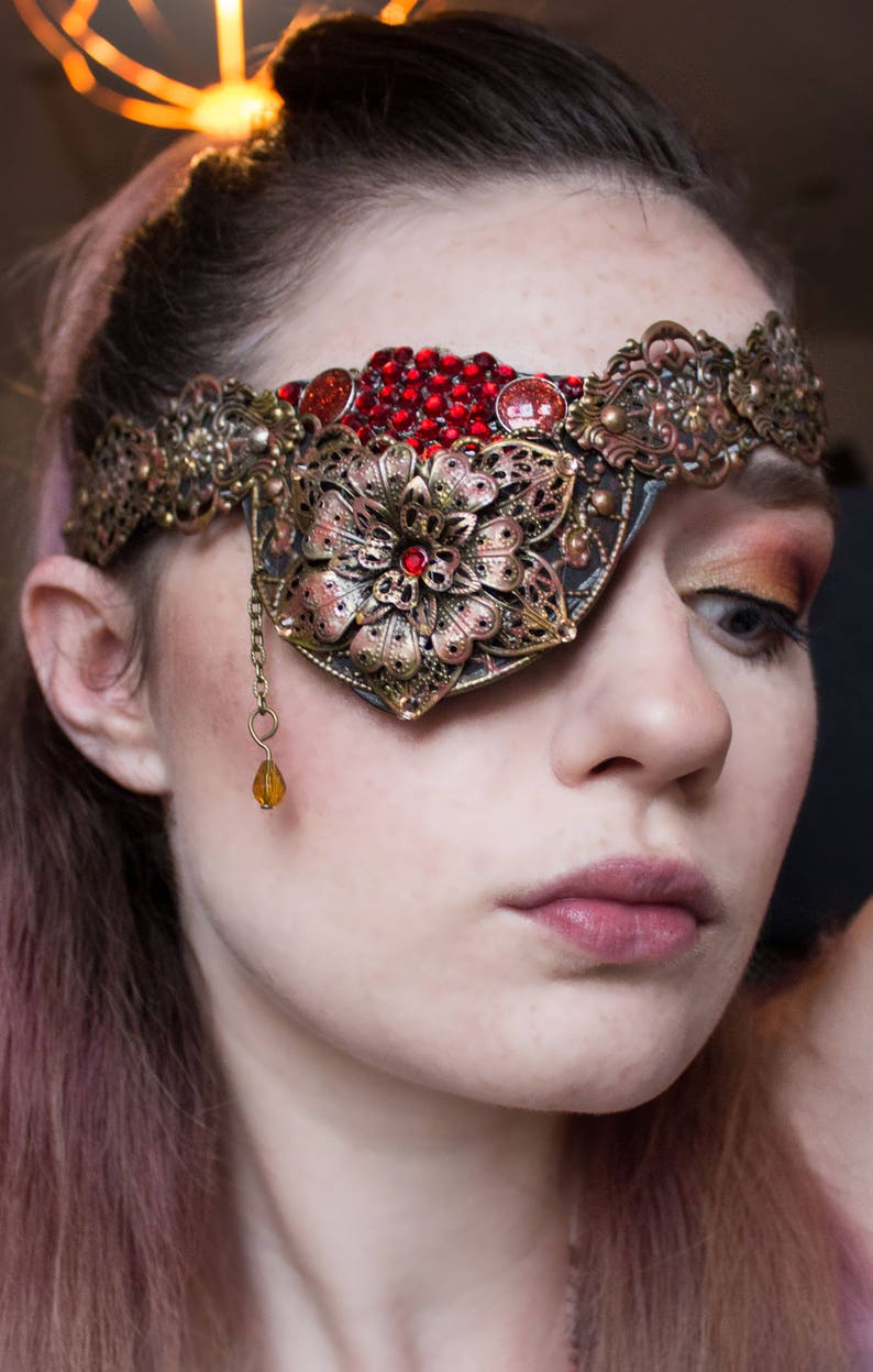 May include: A woman wearing a gold and red eye patch with a jeweled design. The eye patch is attached to a headband with a floral design.