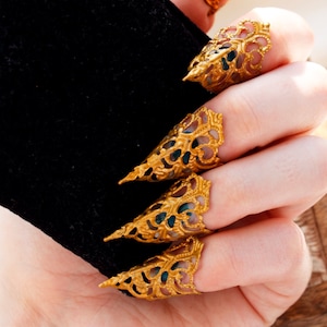 May include: Five gold claw rings with intricate designs. The rings are worn on the fingers of a hand, with the tips of the claws pointing upwards.