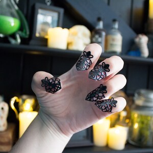 May include: A hand wearing black lace finger rings. The rings are in the shape of a flower with a pointed tip. The hand is in a claw-like position. The background is blurry and shows a shelf with candles and jars.