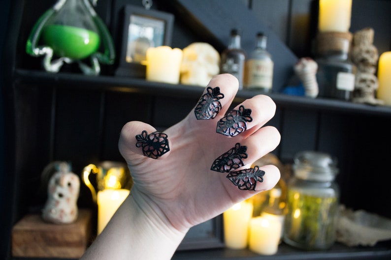 May include: A hand wearing black lace finger rings. The rings are in the shape of a flower with a pointed tip. The hand is in a claw-like position. The background is blurry and shows a shelf with candles and jars.