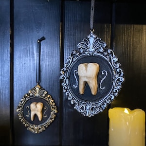 May include: Two black ornate frames with gold accents, each containing a carved wooden tooth. The frames are hanging on a black wooden wall. A lit candle is in the foreground.