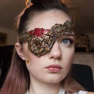 May include: A woman wearing a gold and red eye patch with a jeweled design. The eye patch is attached to a headband with a floral design.