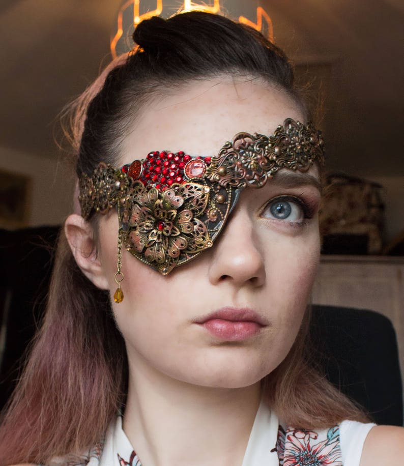May include: A woman wearing a gold and red eye patch with a jeweled design. The eye patch is attached to a headband with a floral design.