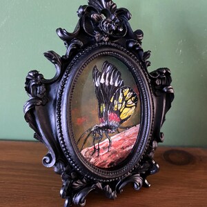 May include: A black ornate oval frame with a painting of a yellow, black, and red butterfly on a brown branch.