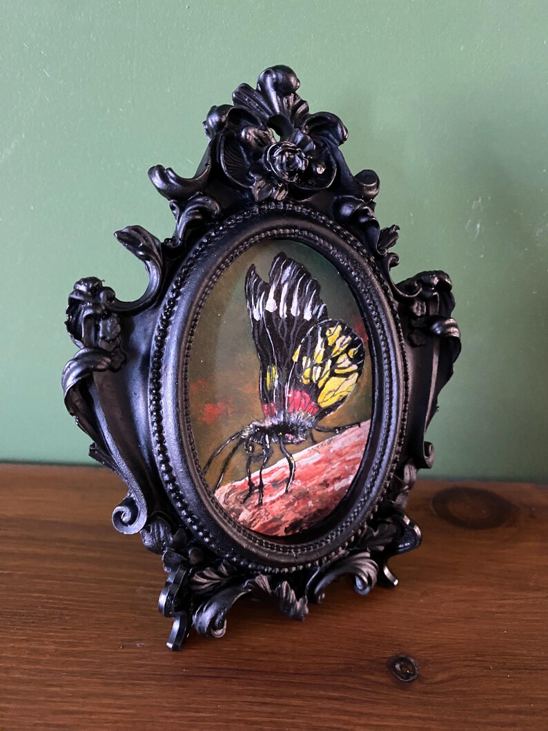 May include: A black ornate oval frame with a painting of a yellow, black, and red butterfly on a brown branch.