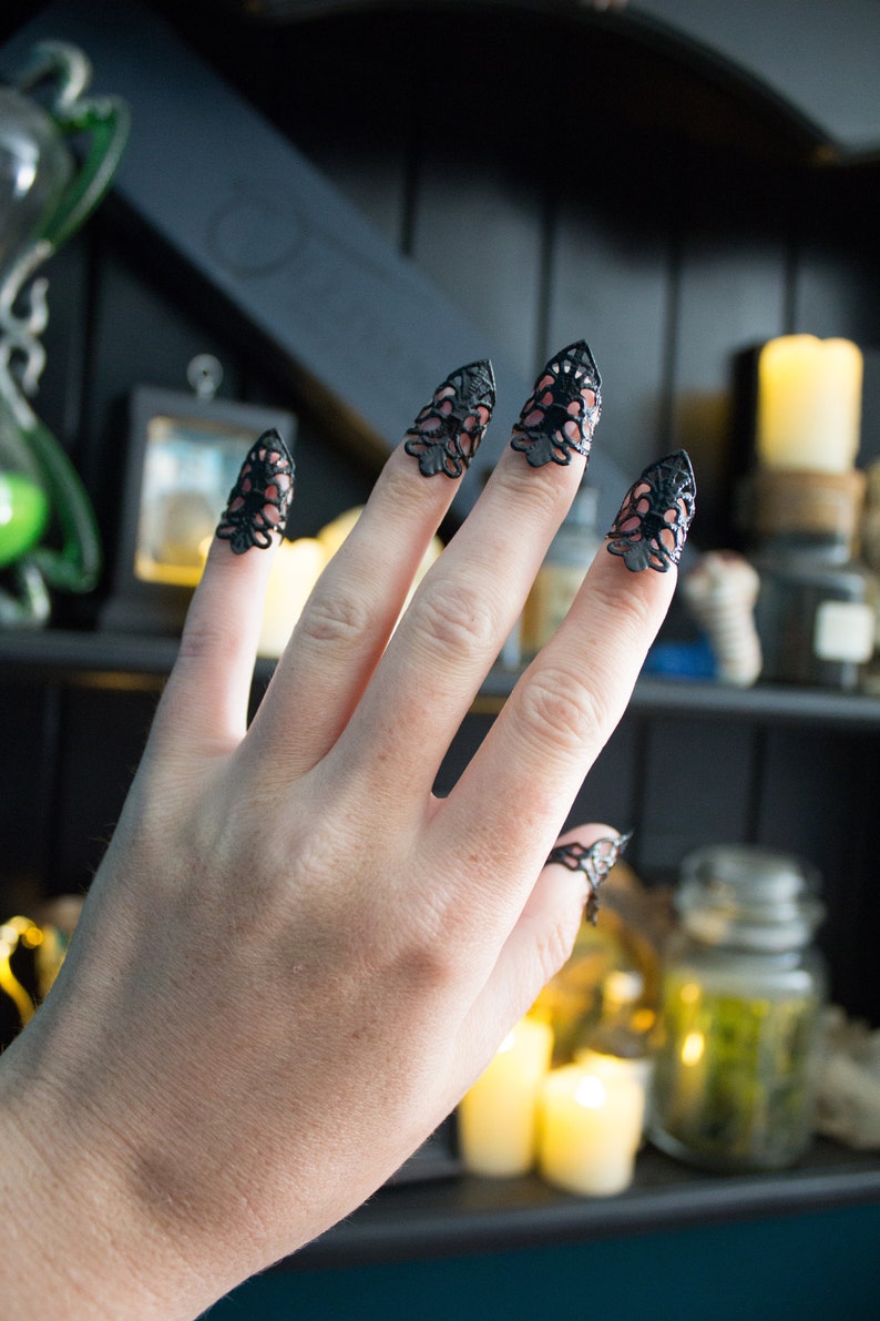 May include: A hand wearing black lace fingertip rings. The rings are in the shape of a pointed claw. The hand is in front of a shelf with candles and jars.