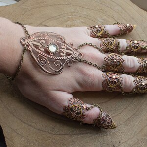Puede incluir: Un conjunto de anillos de dedos adornados, de color dorado, con dise&ntilde;os intrincados y brillantes transparentes. Los anillos est&aacute;n conectados por una cadena y se llevan en varios dedos.
