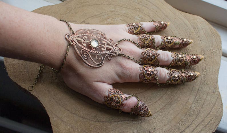 Puede incluir: Un conjunto de anillos de dedos adornados, de color dorado, con dise&ntilde;os intrincados y brillantes transparentes. Los anillos est&aacute;n conectados por una cadena y se llevan en varios dedos.
