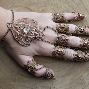 Puede incluir: Un conjunto de anillos de dedo ornamentados, dorados y de cobre con un brazalete grande, decorativo, de filigrana con un cristal transparente. Los anillos est&aacute;n conectados por cadenas.
