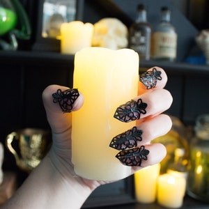 May include: A hand with black lace rings on each finger holds a lit, white pillar candle. The candle is in front of a shelf with various bottles and other decorative items.