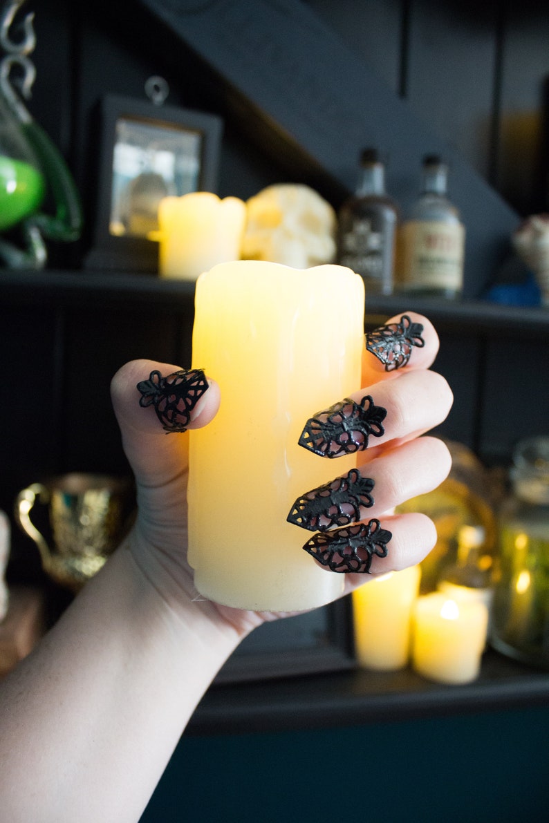 May include: A hand with black lace rings on each finger holds a lit, white pillar candle. The candle is in front of a shelf with various bottles and other decorative items.