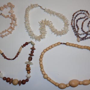 May include: Five necklaces of varying designs and materials are displayed. One is made of light pink stones, another of white shell-like pieces, and a third of brown shells. The remaining two feature wooden beads in shades of brown and tan.