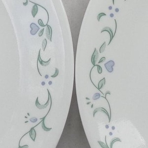 May include: Two white plates with a delicate floral design. The pattern features light green leaves, small blue heart-shaped flowers, and tiny purple dots arranged along the rim. A classic and elegant tableware set.