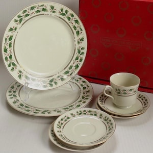 May include: A set of cream-colored ceramic dinnerware featuring a green holly leaf and red berry pattern with gold trim. The set includes two large plates, two saucers, and one teacup, displayed in front of a red box labeled Home for the Holidays.