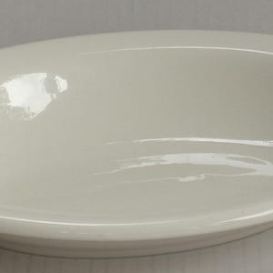 May include: A plain, off-white oval bowl with a smooth, glossy finish and a wide, rounded rim. The bowl features a shallow, concave center and a simple, clean design, showcasing a uniform light cream color throughout its entire surface.