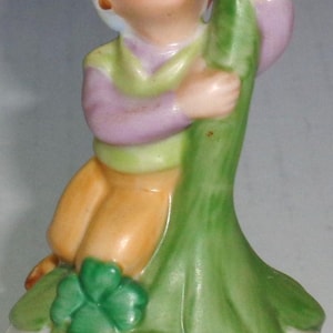 May include: A ceramic figurine of a young person in a green and purple outfit, holding a large green leaf. The figurine has orange pants and a white hat. Three green shamrocks are at the base.