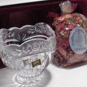 May include: A clear crystal bowl with a scalloped rim featuring etched floral designs. A small bag of dried rose petals with a "Southern Garden Sachet" label is next to the bowl. The bowl has an "ONEIDA Crystal" label.