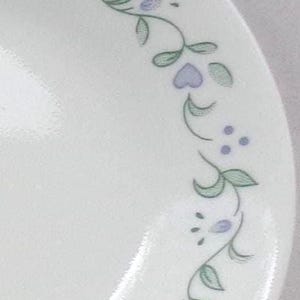 May include: White ceramic plate with a delicate floral design. The border features a repeating pattern of green leaves, lavender hearts, and small purple dots. A simple and elegant design for everyday use.